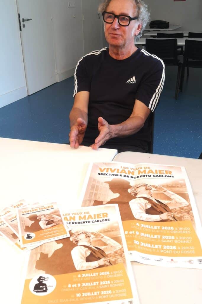 Older man with black glasses sits at a table, gesturing with his hands, with orange Vivian Maier posters spread on the surface.