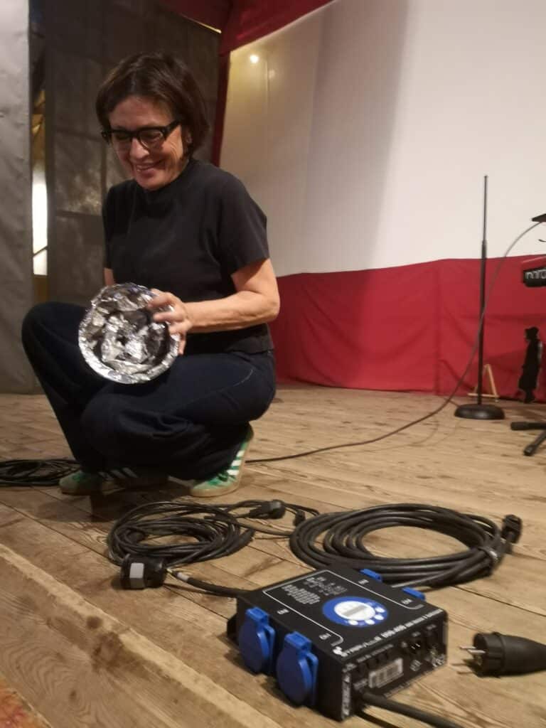 Person crouching on a wooden stage, wearing a black shirt and glasses, holding a crumpled aluminum foil object with both hands.