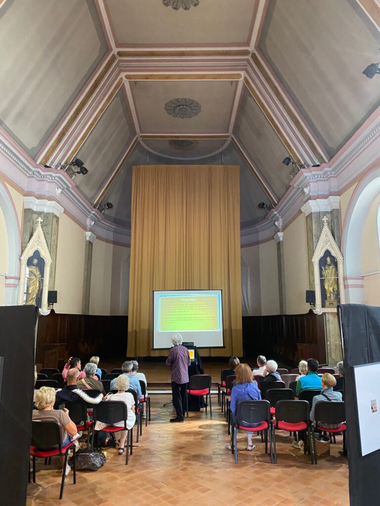 Audience seated in a church hall watching a presentation on a screen in front of a gold curtain stage.
