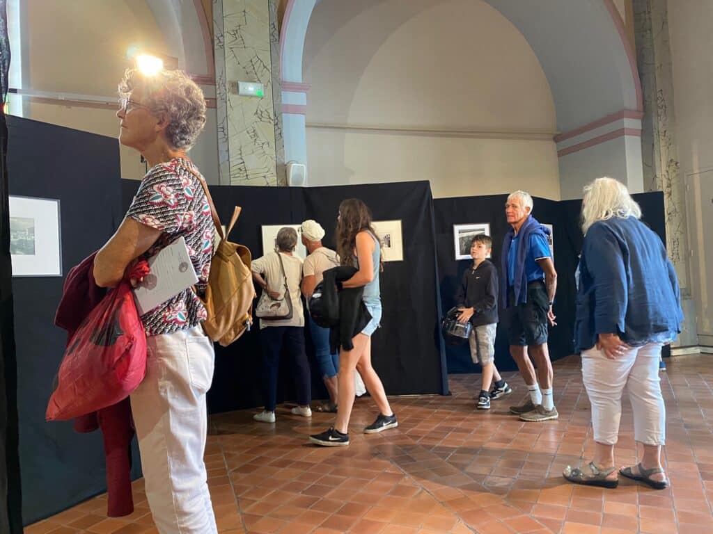 People line up and browse framed artworks displayed on dark partition walls inside a gallery or museum hall.