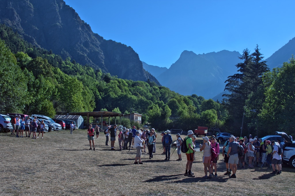 Group of hikers with backpacks gather in a parking area at the base of forested mountains under a clear blue sky.