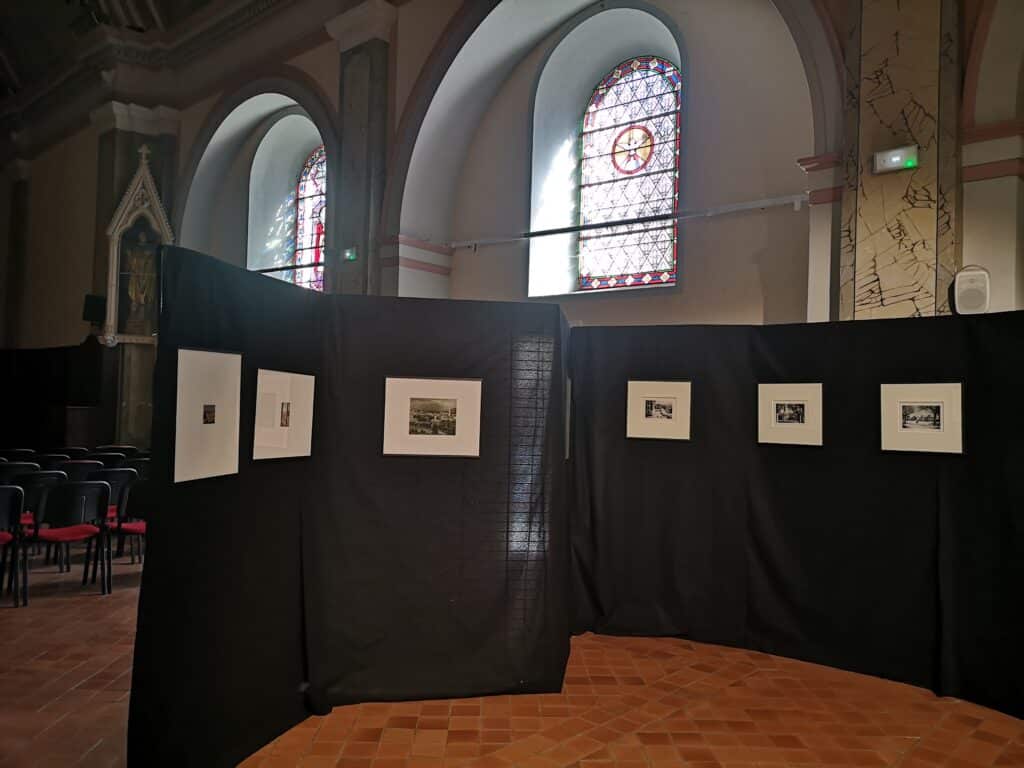 Interior of a church or cathedral with tall arched stained-glass windows and exhibition panels along a partition wall showing framed photos.